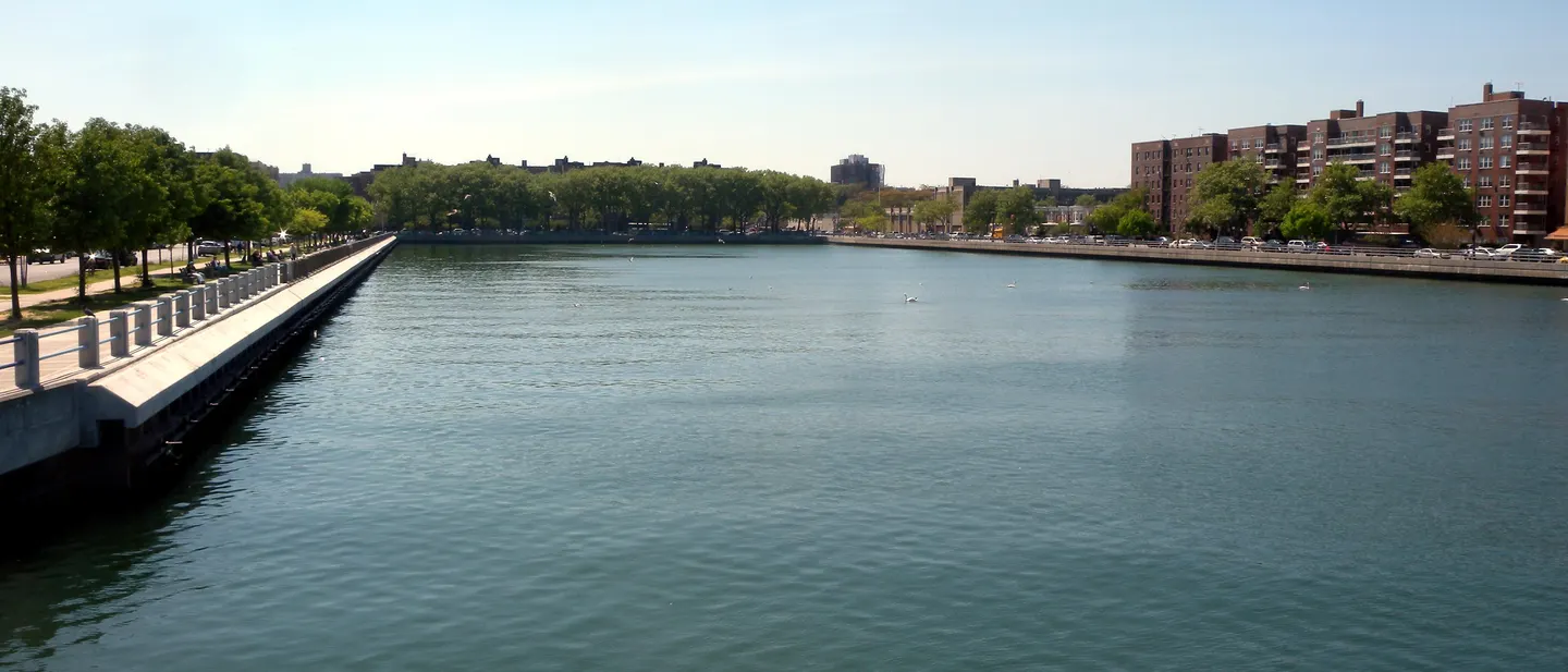 Open water at the head of Sheepshead Bay, a railed promenade running along the near bank and apartment buildings on the far shore.